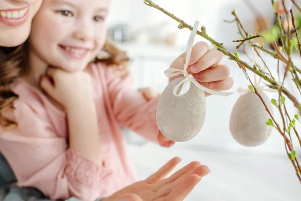 Селективное внимание счастливого ребенка, касающегося декоративного пасхального яйца на ветке ивы рядом с улыбающейся матерью — стоковое фото