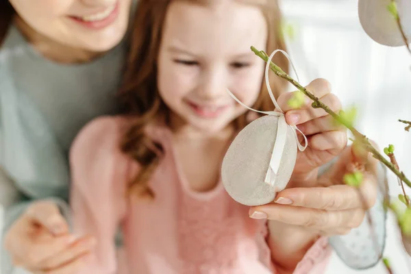 Fuoco selettivo di bambino allegro che tocca l'uovo di Pasqua decorativo su ramo di salice vicino a madre sorridente — Foto stock