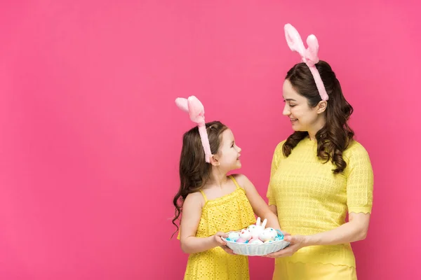 Happy mother and daughter in bunny ears holding easter eggs and looking at each other isolated on pink — Stock Photo