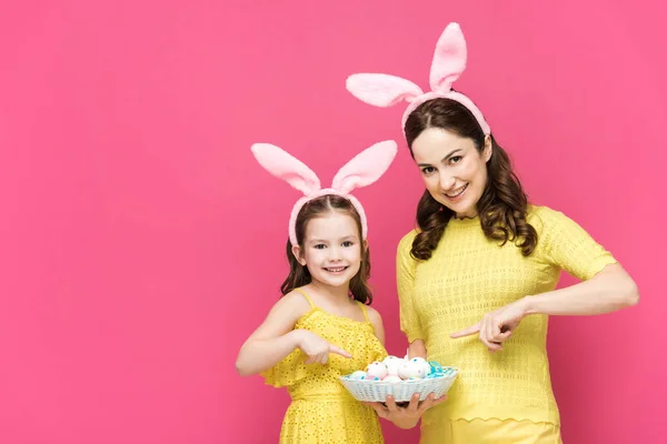 Feliz madre e hija en orejas de conejo apuntando con los dedos a los huevos de Pascua aislados en rosa - foto de stock