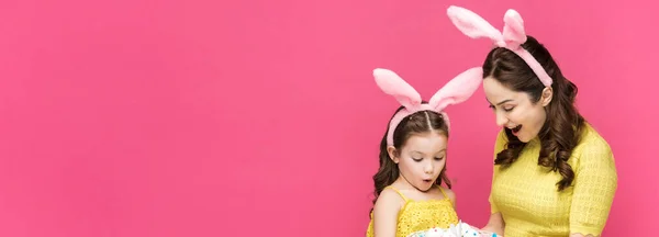 Plan panoramique de mère et fille surprise dans des oreilles de lapin regardant des œufs de Pâques isolés sur rose — Photo de stock