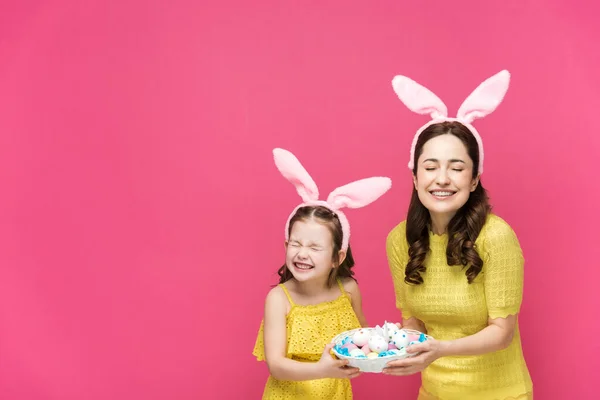 Mãe feliz e filha em orelhas de coelho segurando ovos de Páscoa e rindo isolado em rosa — Fotografia de Stock