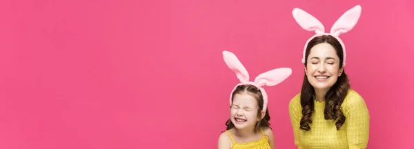 Plan panoramique de mère et fille heureuses dans des oreilles de lapin tenant des œufs de Pâques et riant isolé sur rose — Photo de stock