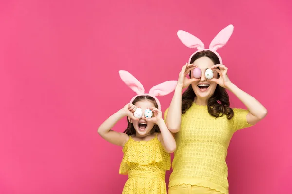 Excited mother and daughter in bunny ears covering eyes with easter eggs isolated on pink — Stock Photo