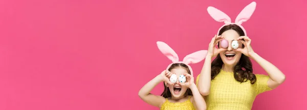 Tiro panorâmico de mãe excitada e filha em orelhas de coelho cobrindo olhos com ovos de Páscoa isolados em rosa — Fotografia de Stock
