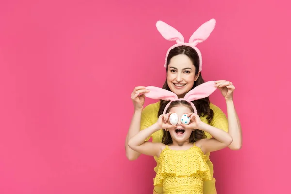 Felice madre toccare coniglietto orecchie di figlia che copre gli occhi con uova di Pasqua isolate su rosa — Foto stock