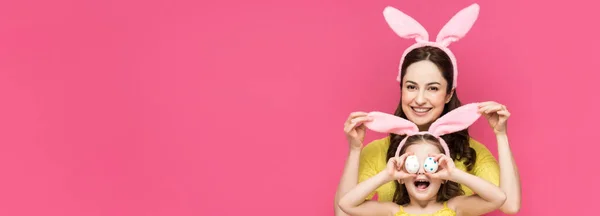 Panoramic shot of happy mother touching bunny ears of daughter covering eyes with easter eggs isolated on pink — Stock Photo