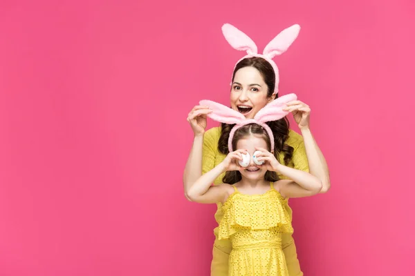 Mãe excitada tocando orelhas de coelho da filha cobrindo olhos com ovos de Páscoa isolados em rosa — Fotografia de Stock