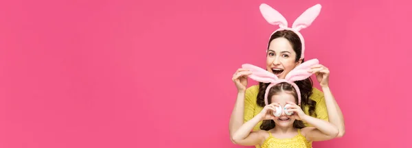 Панорамный снимок взволнованной матери, трогающей кроличьи уши дочери, закрывающей глаза пасхальными яйцами, изолированными на розовом — стоковое фото