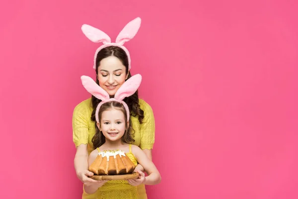 Happy mother in bunny ears near daughter with easter cake isolated on pink — Stock Photo