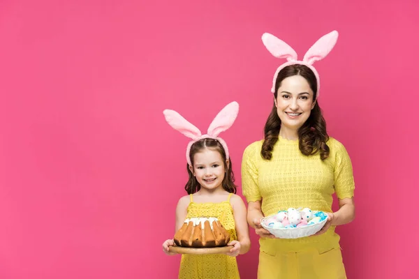 Mère heureuse dans les oreilles de lapin tenant des œufs de poulet peints près de la fille avec gâteau de Pâques isolé sur rose — Photo de stock