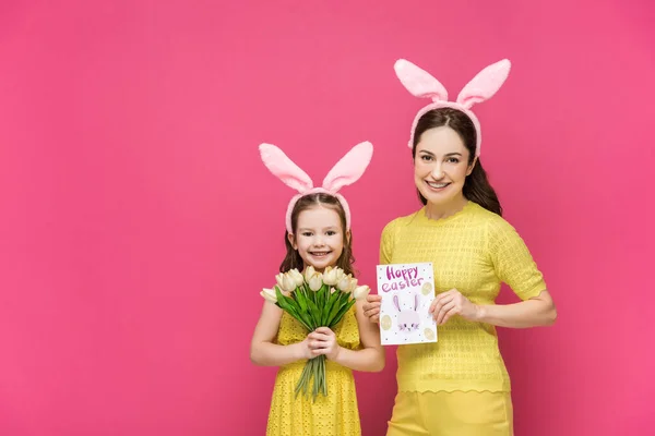 Happy mother in bunny ears holding greeting card with happy easter lettering near daughter with tulips isolated on pink — Stock Photo