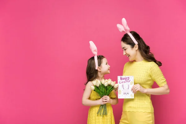 Fröhliche Mutter in Hasenohren hält Grußkarte mit fröhlichem Osteraufdruck in der Nähe der Tochter mit Tulpen auf rosa — Stockfoto