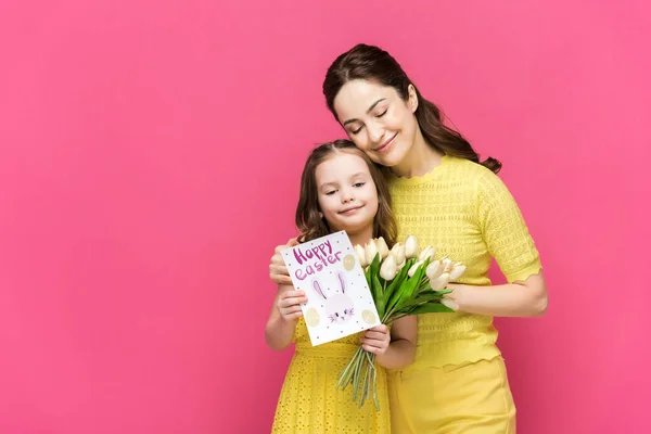 Fröhliche Mutter hält Glückwunschkarte mit frohem Osteraufdruck und umarmt Tochter mit Tulpen isoliert auf rosa — Stockfoto