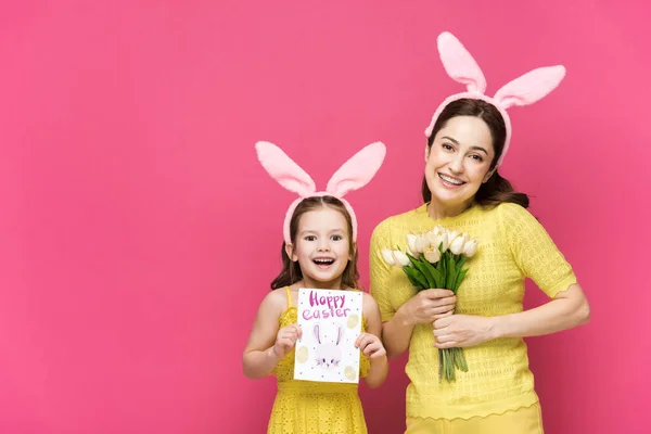 Веселая мать в кроличьих ушах держит поздравительную открытку со счастливой пасхальной надписью рядом с ребенком с тюльпанами изолированы на розовый — стоковое фото
