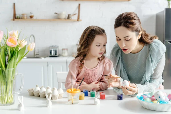 Criança feliz e mãe pintando ovos de Páscoa perto de tulipas e coelhos decorativos — Fotografia de Stock