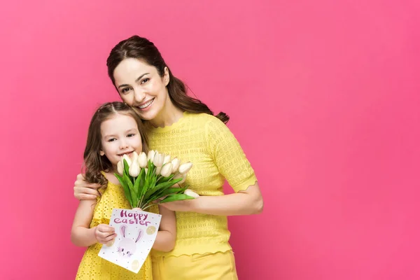 Allegra madre in possesso di biglietto di auguri con felice lettering pasquale e abbraccio bambino con tulipani isolati su rosa — Foto stock