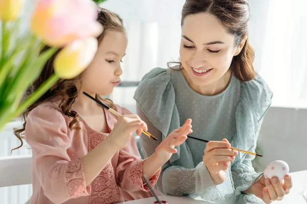 Selective focus of happy mother and daughter painting chicken egg near tulips — Stock Photo