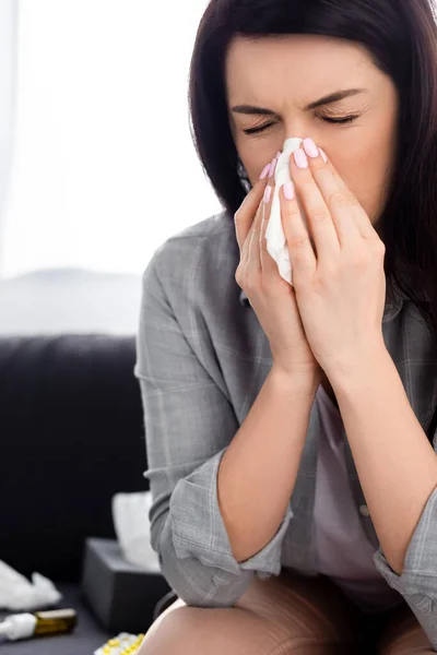 Allergische Frau mit geschlossenen Augen niest in Serviette, während sie auf dem Sofa sitzt — Stockfoto