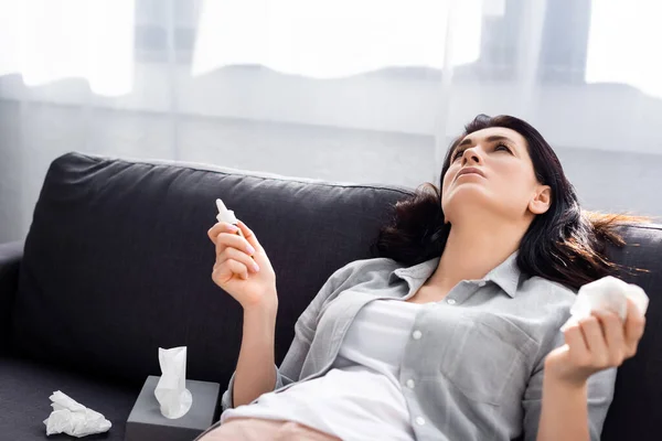 Allergic woman holding napkin and nasal spray while lying on sofa — Stock Photo