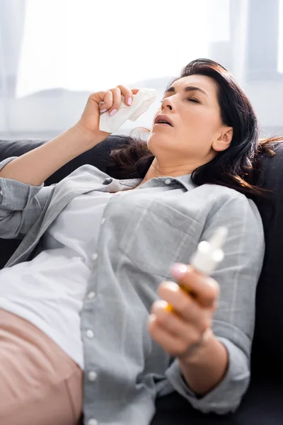 Selective focus of allergic woman holding napkin and nasal spray while lying on sofa — Stock Photo