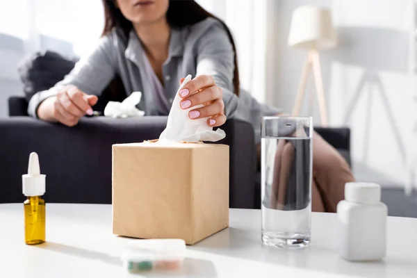 Vista recortada de la mujer alérgica que toma tejido cerca del vaso de agua, botella con pastillas y aerosol nasal - foto de stock