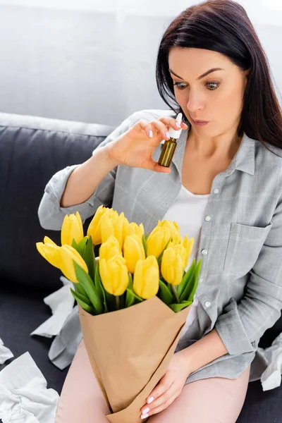 Woman with pollen allergy sitting on sofa with flowers and holding nasal spray — Stock Photo