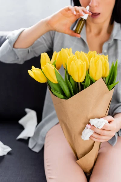 Cropped view of woman with pollen allergy sitting on sofa with flowers and holding nasal spray — Stock Photo