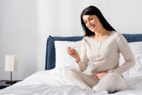 Счастливая женщина с тестом на беременность и сидящая на кровати — стоковое фото