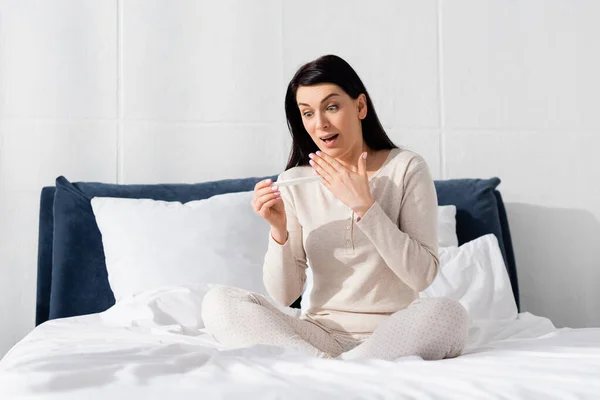 Здивована жінка тримає тест на вагітність і сидить на ліжку — стокове фото