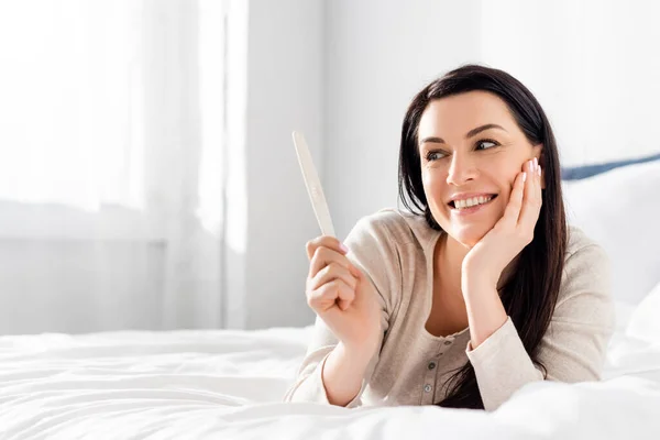 Femme enceinte souriant et tenant test de grossesse tout en étant couché sur le lit — Photo de stock