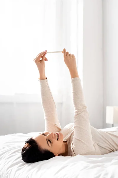 Вагітна жінка посміхається і дивиться на тест на вагітність, лежачи на ліжку — стокове фото
