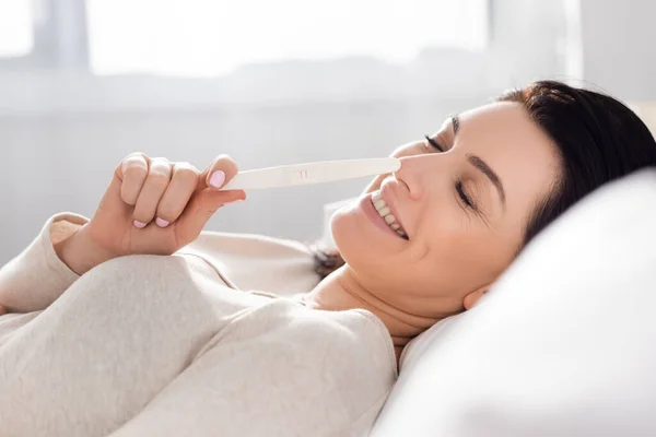 Счастливая женщина смотрит тест на беременность, лежа на кровати — стоковое фото