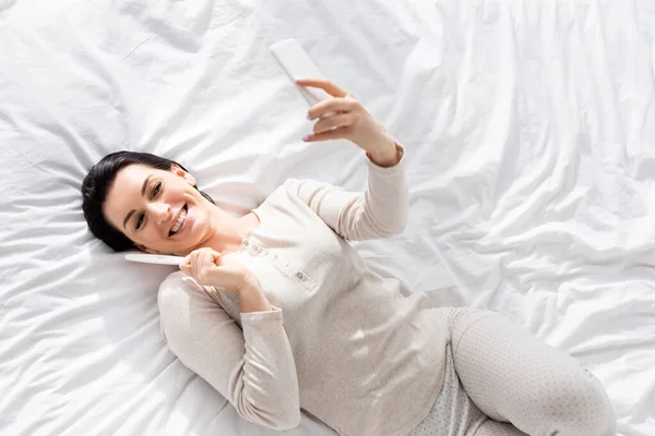 Vista dall'alto della donna felice che prende selfie con test di gravidanza mentre si sdraia sul letto — Foto stock