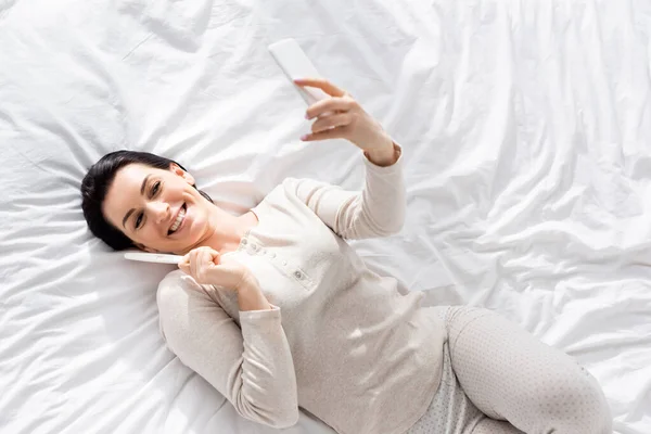 Vista superior de la mujer feliz tomando selfie con prueba de embarazo mientras está acostado en la cama — Stock Photo
