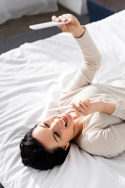 Над головой вид счастливой женщины, делающей селфи с тестом на беременность, лежащей на кровати — стоковое фото