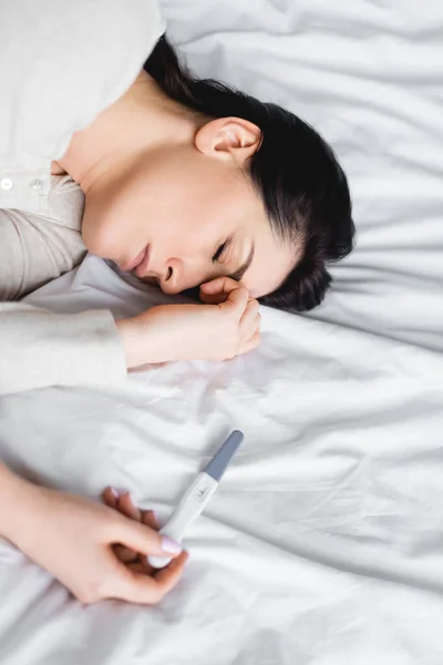 Mulher deprimida com os olhos fechados deitada na cama perto do teste de gravidez — Fotografia de Stock