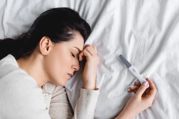 Vista superior de la mujer deprimida con los ojos cerrados acostados en la cama cerca de la prueba de embarazo - foto de stock
