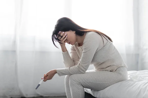 Traurige Frau sitzt auf Bett und hält Schwangerschaftstest mit negativem Ergebnis — Stockfoto