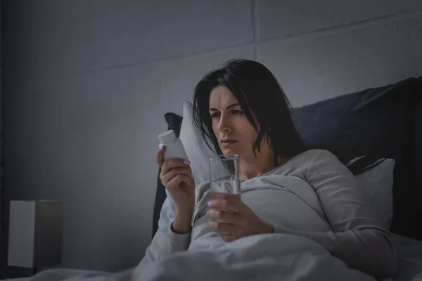 Mujer con trastorno del sueño sosteniendo vaso de agua y botella con pastillas para dormir en el dormitorio - foto de stock