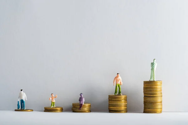 Figuras de pessoas de plástico em moedas de ouro empilhadas na superfície branca em fundo cinza, conceito de igualdade financeira — Fotografia de Stock