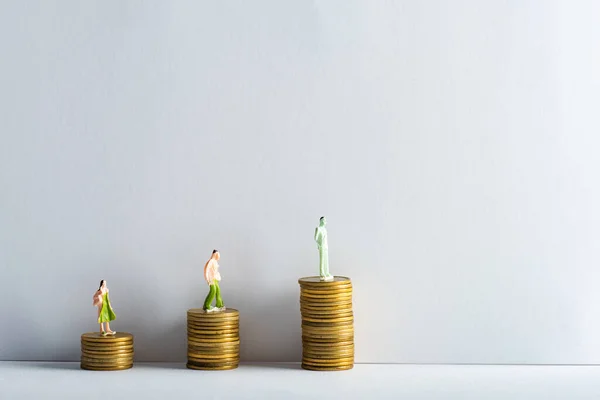 Três pessoas figuras em moedas de ouro empilhadas na superfície branca em fundo cinza, conceito de igualdade financeira — Fotografia de Stock