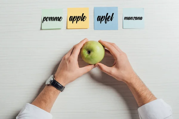 Vue recadrée de l'homme d'affaires tenant une pomme sur la table avec des notes collantes avec traduction de pomme — Photo de stock