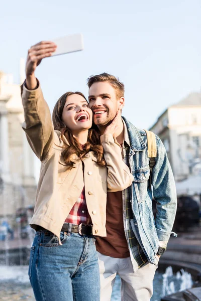 Selective focus of excited and happy couple taking selfie near fountain in city — Stock Photo