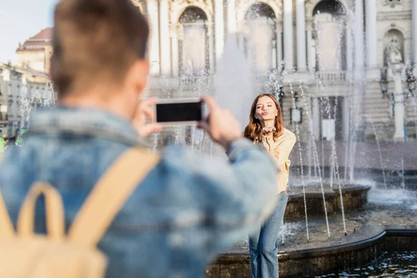 Selective focus of man taking picture with smartphone of woman blowing kiss near fountain in city — Stock Photo