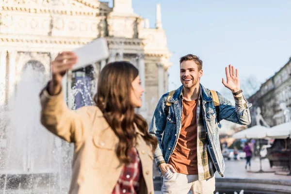 Selective focus of woman with boyfriend waving hand taking selfie with smartphone near fountain in city — Stock Photo