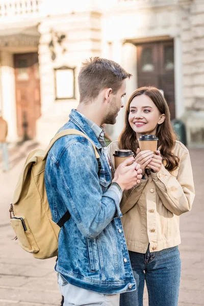 Хлопець і дівчина тримають паперові чашки кави зі згорнутими руками, дивлячись один на одного і посміхаючись в місті — стокове фото