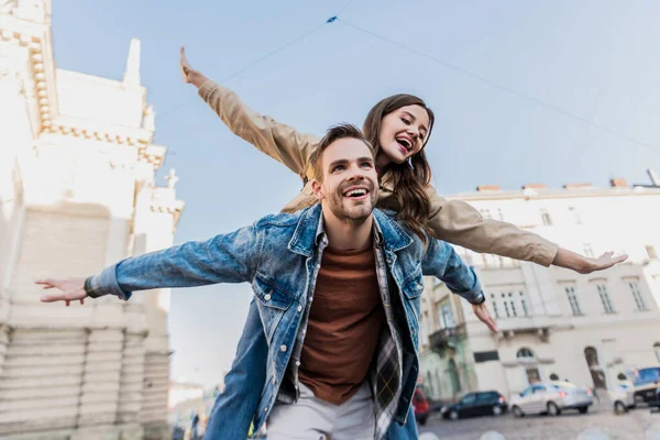 Low angle view of man smiling and piggybacking excited girl with open arms in city — Stock Photo