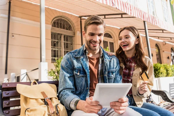 Селективный фокус пары улыбающейся и держащей кредитную карту и цифровой планшет на скамейке в городе — стоковое фото