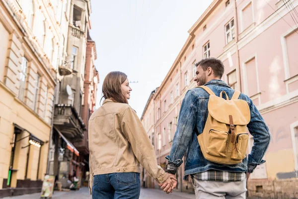 Visão traseira do casal olhando um para o outro e de mãos dadas com mochila na cidade — Fotografia de Stock